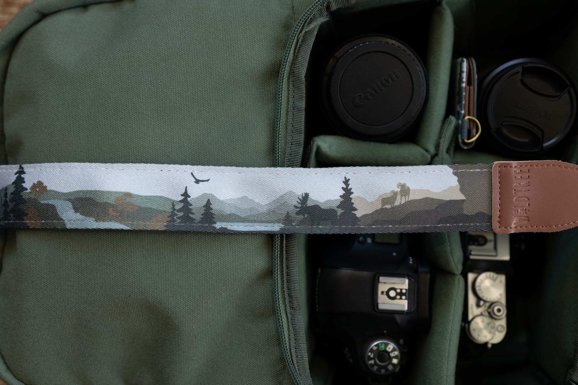 Close-up of a camera strap featuring a stylized nature illustration with pine trees, a moose silhouette, and an eagle flying over a mountain landscape, attached to a brown leather end piece with the logo "WILDTREE"