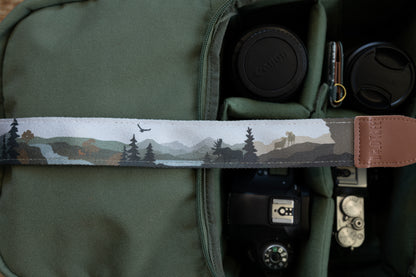 Close-up of a camera strap featuring a stylized nature illustration with pine trees, a moose silhouette, and an eagle flying over a mountain landscape, attached to a brown leather end piece with the logo "WILDTREE"