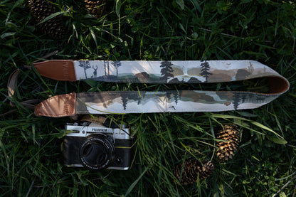 Two camera straps laid out on grass next to a camera; both feature the same artistic wilderness print of forests, mountains, and animals, with branded "WILDTREE" leather ends.