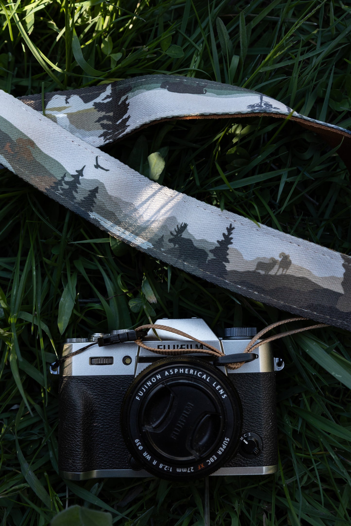 The camera strap draped over a camera body on grass, showing the detailed print of a moose, a flock of birds, and forested mountain terrain.