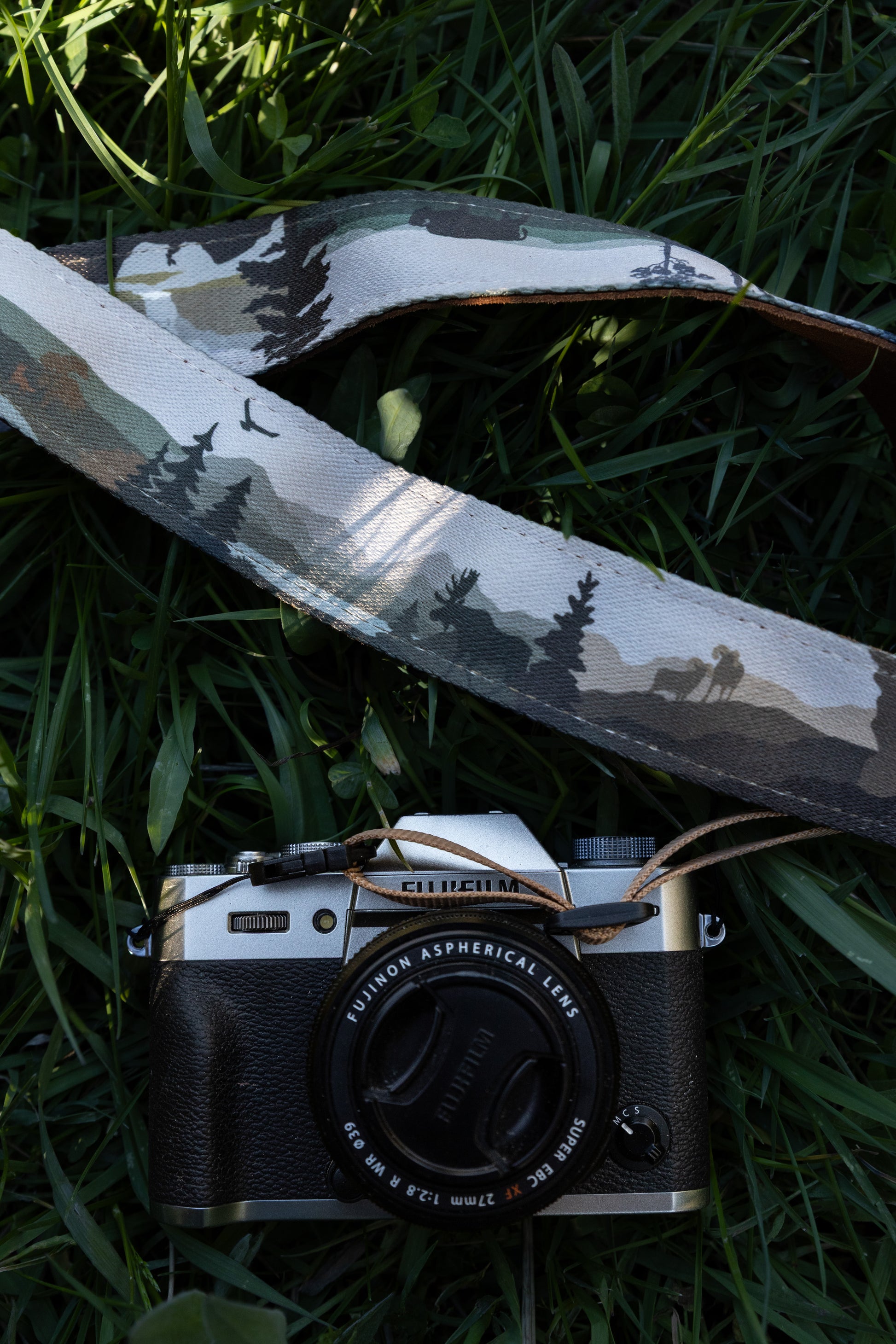 The camera strap draped over a camera body on grass, showing the detailed print of a moose, a flock of birds, and forested mountain terrain.