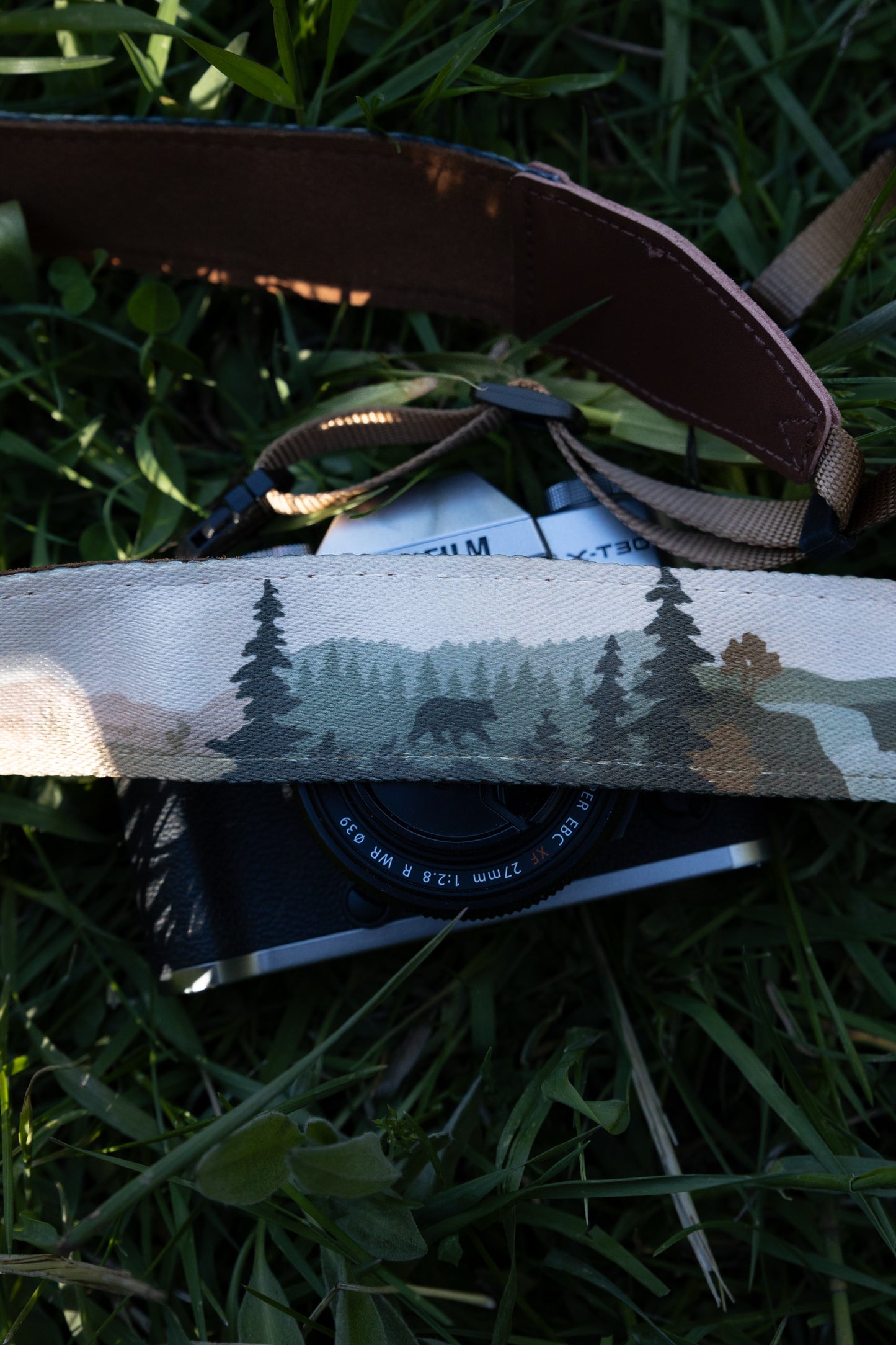 The camera strap resting on grass, featuring a close-up of the nature design with a black bear silhouette surrounded by evergreen trees and a landscape background.