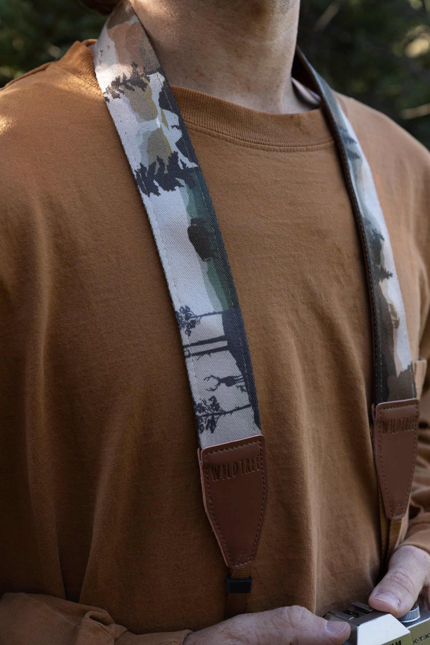 A person wearing the camera strap, which features a repeating scenic print of wilderness landscapes, pine forests, and wildlife, finished with branded "WILDTREE" brown leather connectors.