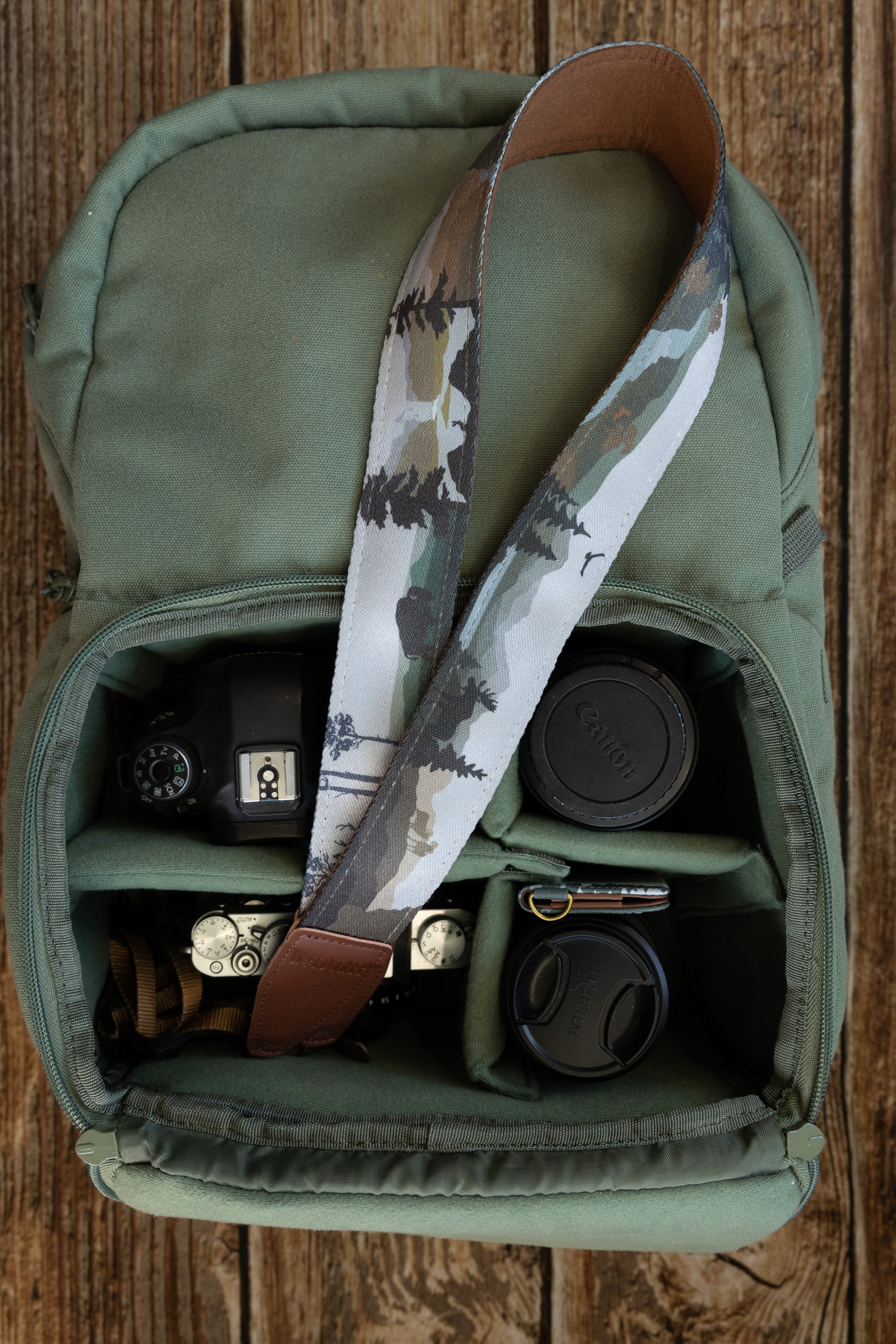 A camera strap draped inside a gear bag, showcasing the continuous nature-themed print that includes pine trees, mountains, and wildlife silhouettes.