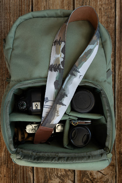 A camera strap draped inside a gear bag, showcasing the continuous nature-themed print that includes pine trees, mountains, and wildlife silhouettes.