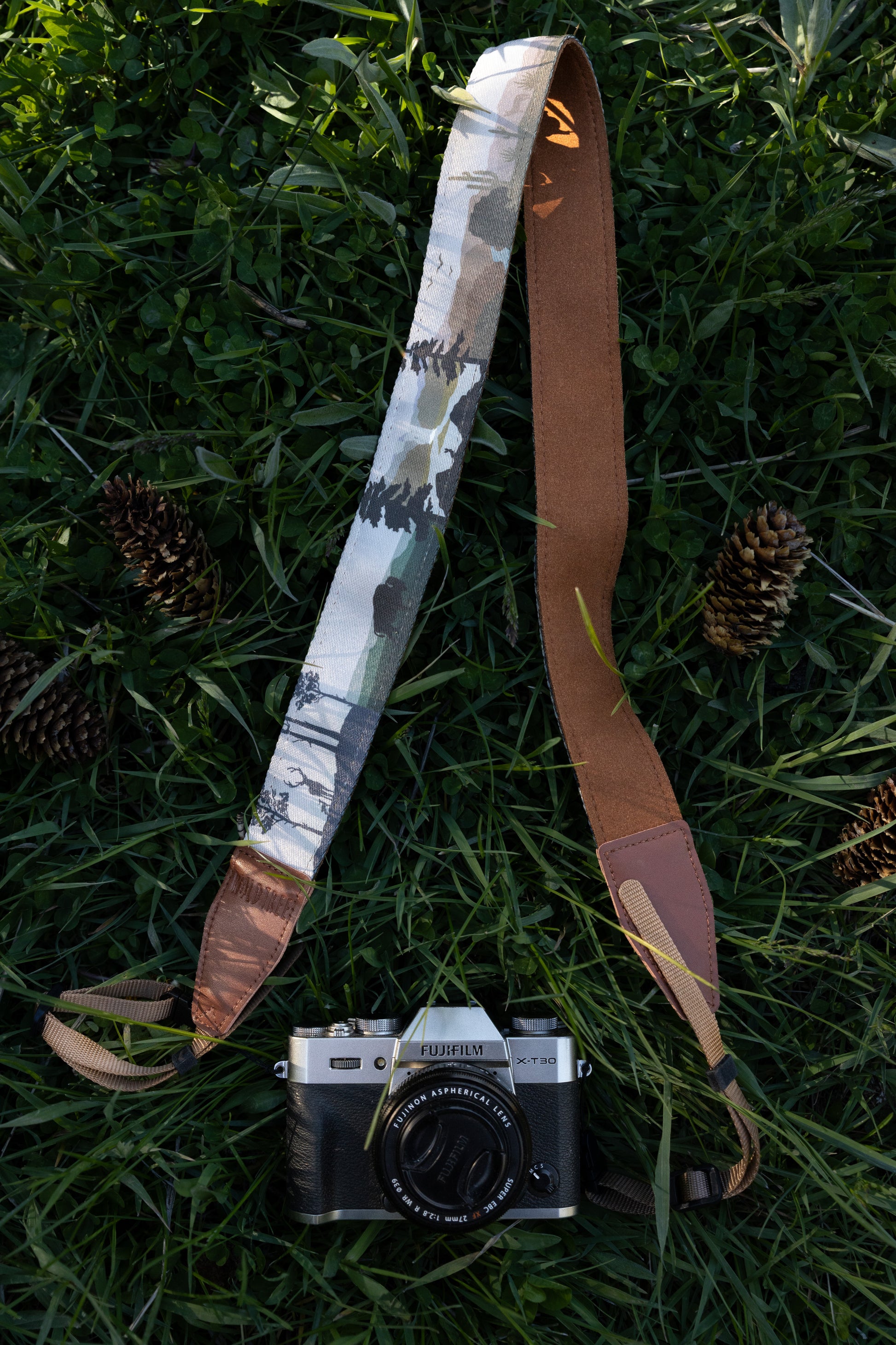 A camera strap with a printed nature design featuring forests and mountain silhouettes attached to a camera, shown resting on a grassy surface with pinecones.