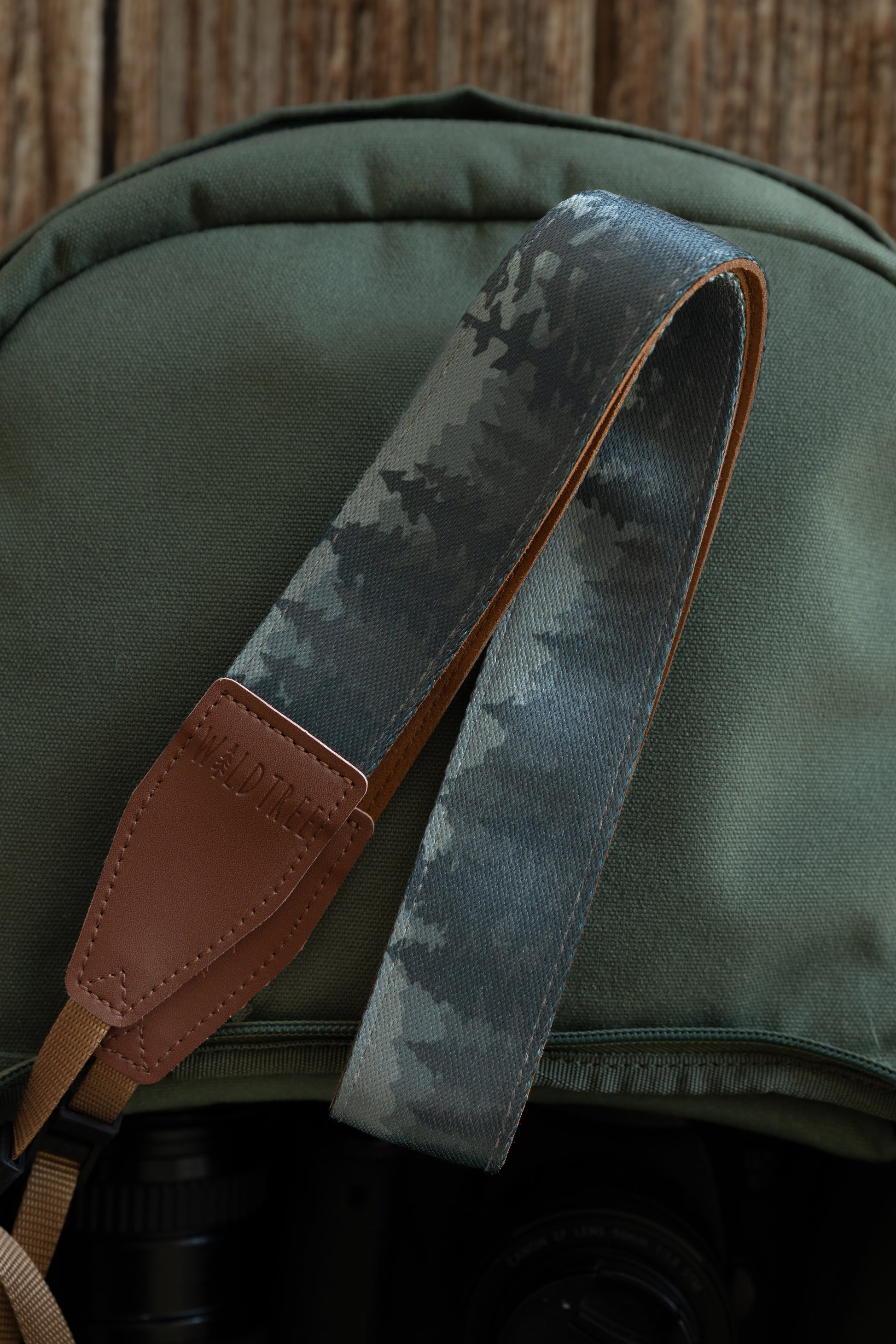 A close-up, vertical shot of a foggy forest camera strap draped over a green canvas backpack. The strap features a dark teal and grey evergreen tree print with brown vegan leather ends.