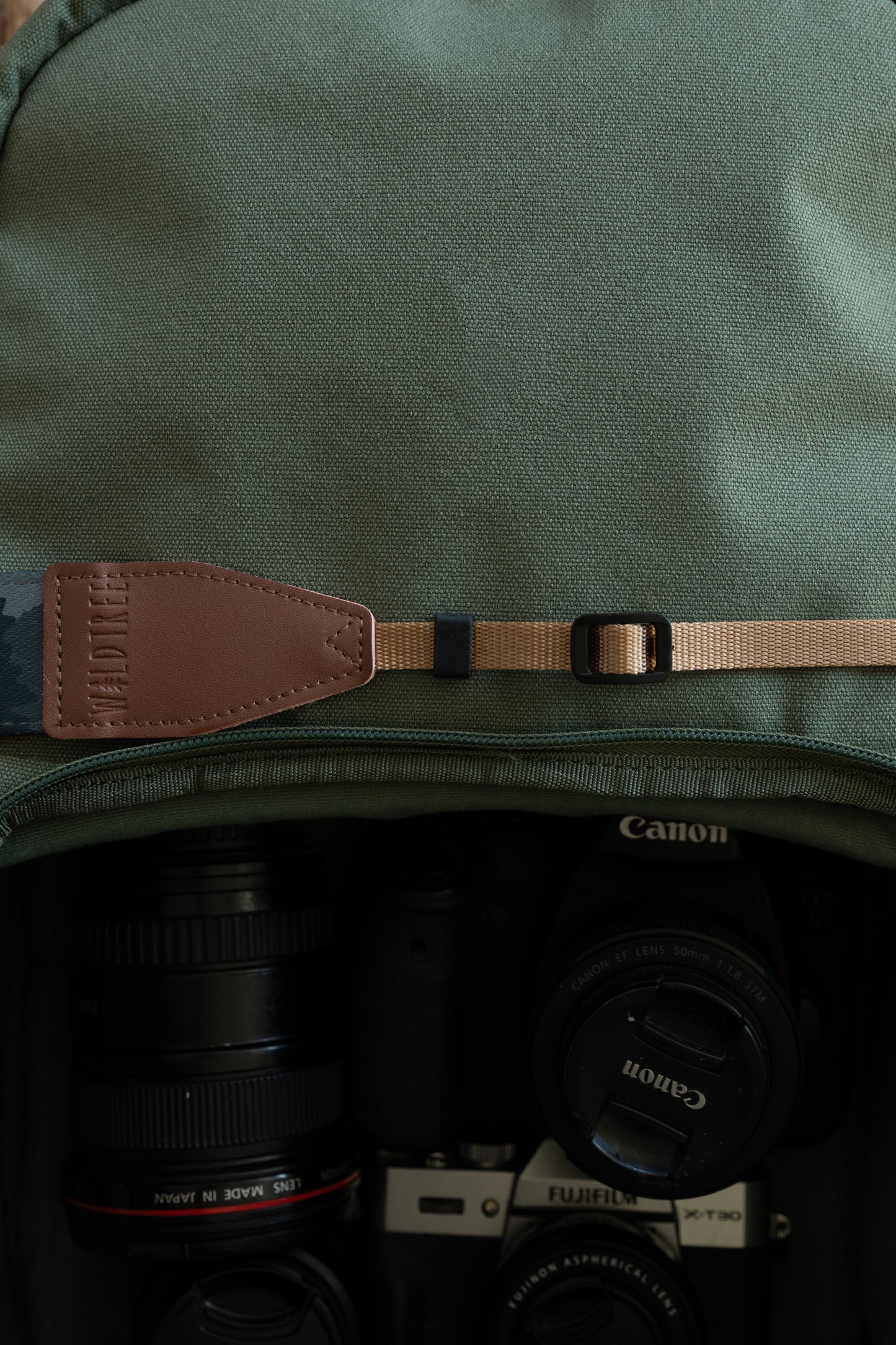Detail shot focusing on the adjustable tan nylon webbing and a brown vegan leather connector embossed with the "WILDTREE" logo, resting against a green camera bag.