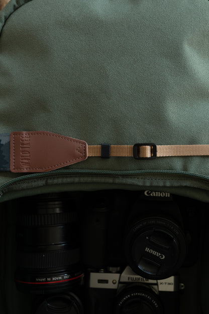 Detail shot focusing on the adjustable tan nylon webbing and a brown vegan leather connector embossed with the "WILDTREE" logo, resting against a green camera bag.