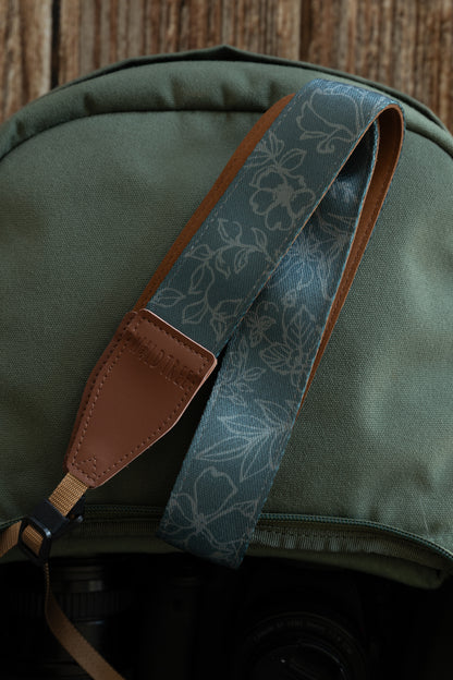 A close-up view of the Wildtree Shadow Garden camera strap folded diagonally over a green backpack. The strap displays a teal-colored fabric with a delicate, etched floral design and a brown leather connector at the end. A tan nylon adjustment webbing with a black plastic buckle extends from the leather piece. The background shows the texture of the green canvas bag and a rustic wooden surface.
