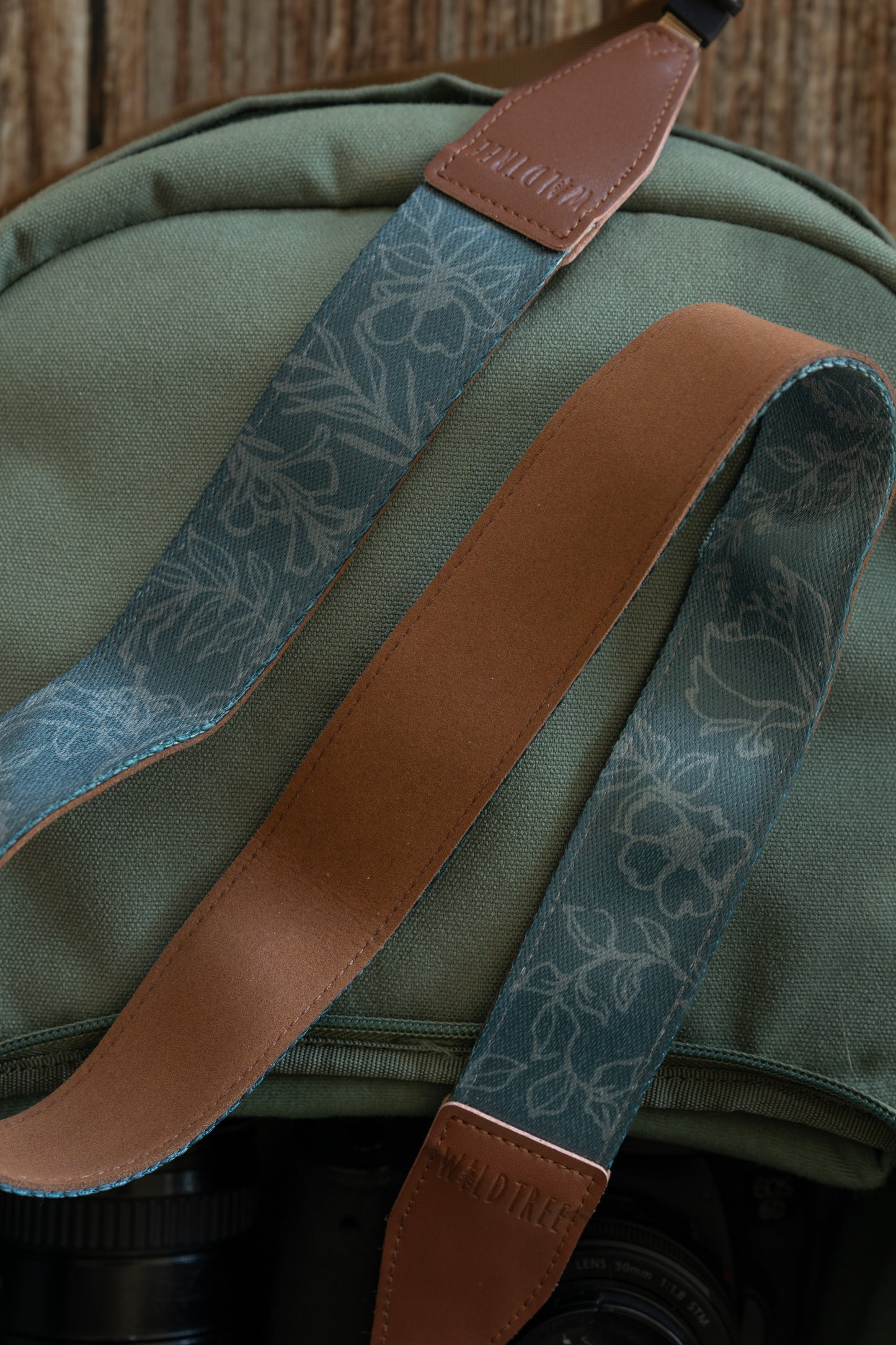 An extreme close-up highlighting the contrast between the two sides of the camera strap. One section shows the teal fabric with its intricate grey floral outlines, while the overlapping section reveals the smooth, solid brown underside of the strap. The embossed "WILDTREE" leather end piece is visible at the top and bottom of the frame against the green canvas background.