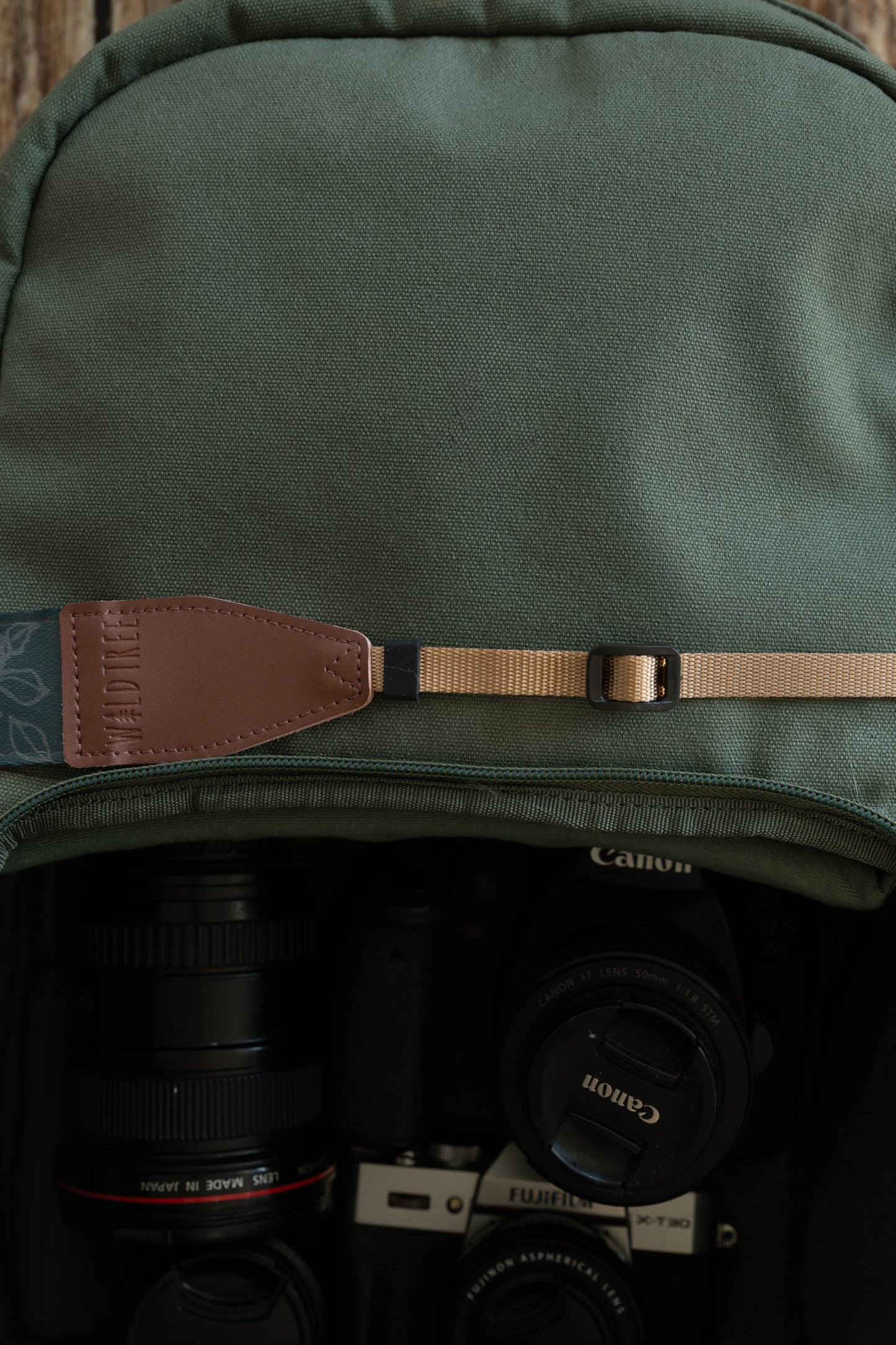 A detailed shot focusing on the attachment point of the camera strap. It shows a brown leather end piece with the "WILDTREE" logo stitched to a tan nylon webbing strap. A black plastic slider buckle is visible on the tan webbing, used for adjusting the length. The strap sits against a green backpack, with camera gear visible in the blurred compartment below.