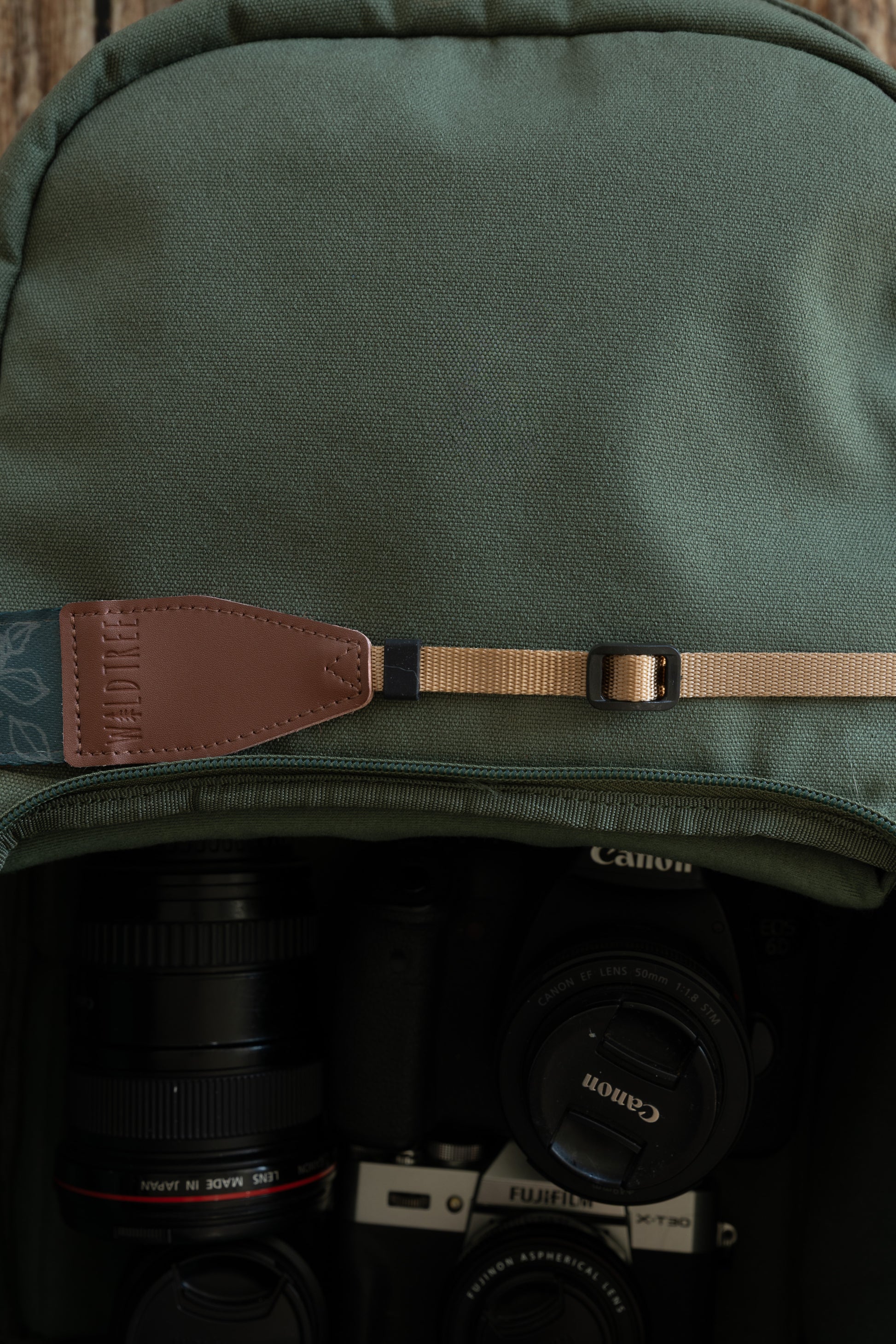 A detailed shot focusing on the attachment point of the camera strap. It shows a brown leather end piece with the "WILDTREE" logo stitched to a tan nylon webbing strap. A black plastic slider buckle is visible on the tan webbing, used for adjusting the length. The strap sits against a green backpack, with camera gear visible in the blurred compartment below.