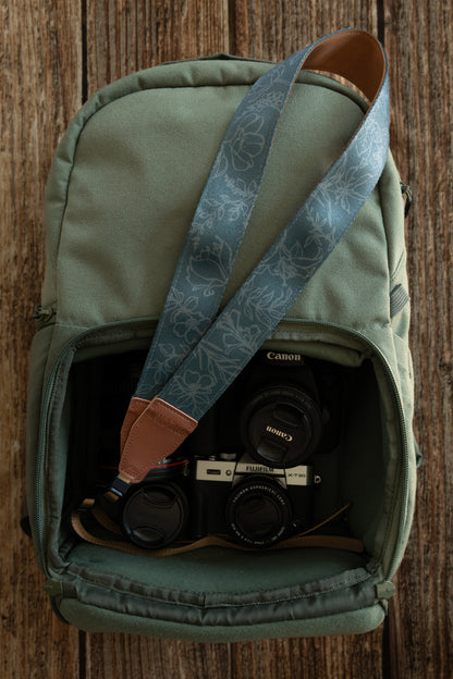 A high-angle shot of a green canvas camera backpack lying on a wooden surface. A teal floral-patterned camera strap is draped loosely over the bag, with one end tucked into the main compartment. Inside the bag, a Fujifilm X-T30 camera and a Canon DSLR with a 50mm lens are neatly packed among other photography equipment.