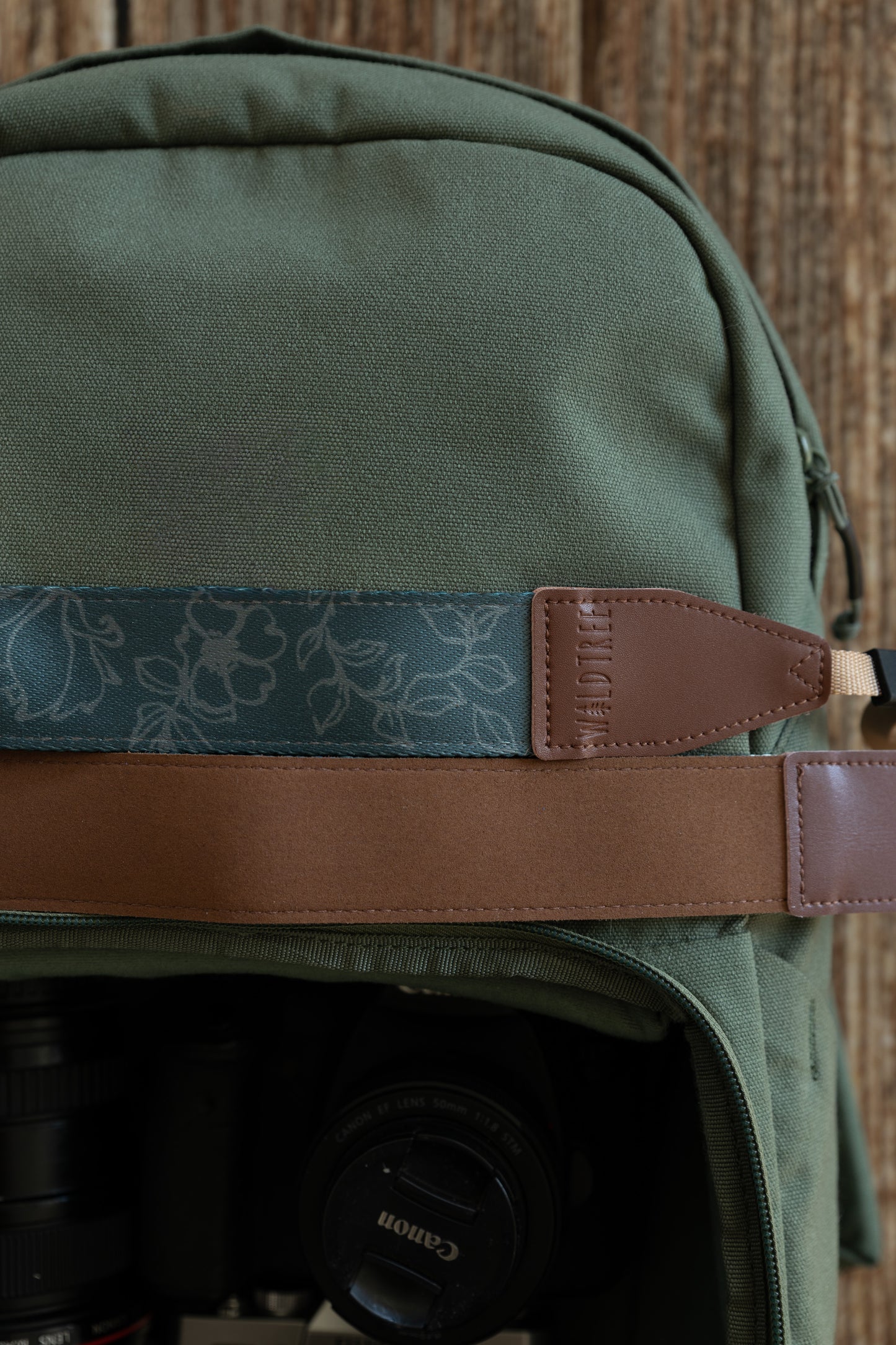 A close-up, top-down shot showing two camera straps resting horizontally across a green canvas backpack. The top strap features a teal fabric with a light grey floral "Shadow Garden" pattern and a brown leather end piece embossed with the "WILDTREE" logo. The bottom strap is a solid, smooth brown material. Below the straps, the open compartment of the backpack reveals a Canon camera lens and body.