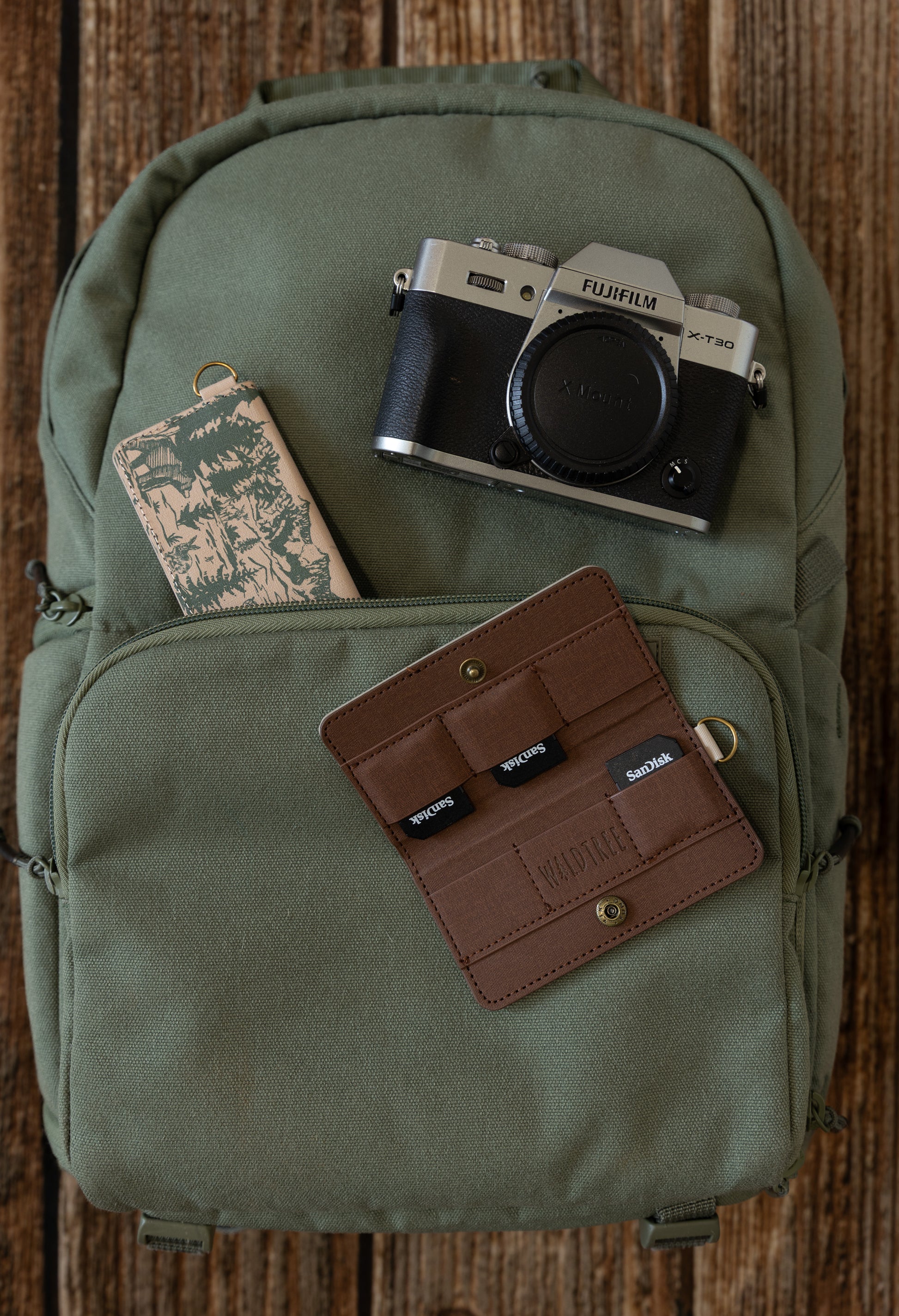 A flat lay on an olive green backpack featuring a Fujifilm camera and two WILDTREE SD card holders—one open showing organized cards and one closed showing the mountain peak design.