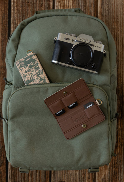 A flat lay on an olive green backpack featuring a Fujifilm camera and two WILDTREE SD card holders—one open showing organized cards and one closed showing the mountain peak design.