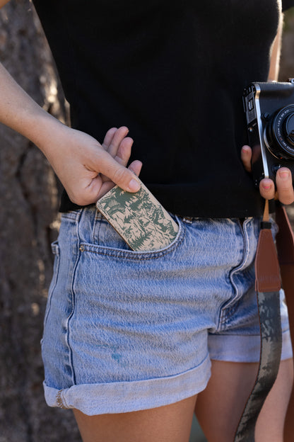 A person sliding a slim, tan and green woodland-patterned SD card holder into the front pocket of blue denim shorts to demonstrate portability.