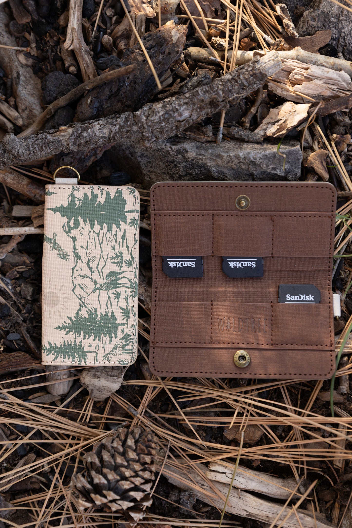An open and closed WILDTREE SD card holder on a forest floor. The brown interior displays multiple SD cards tucked into secure slots next to the debossed WILDTREE logo.