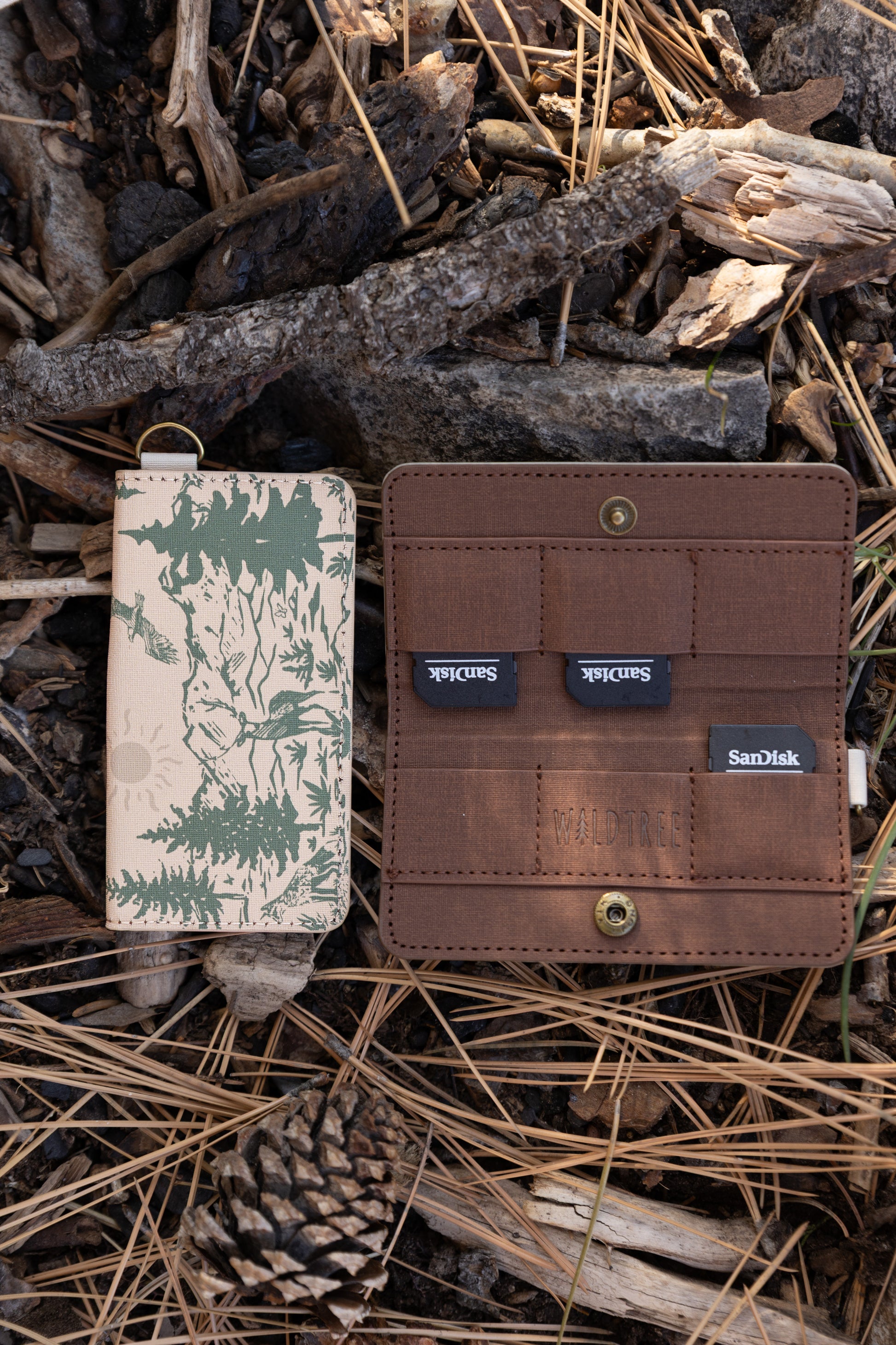 An open and closed WILDTREE SD card holder on a forest floor. The brown interior displays multiple SD cards tucked into secure slots next to the debossed WILDTREE logo.