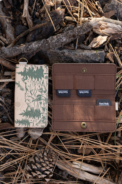 An open and closed WILDTREE SD card holder on a forest floor. The brown interior displays multiple SD cards tucked into secure slots next to the debossed WILDTREE logo.