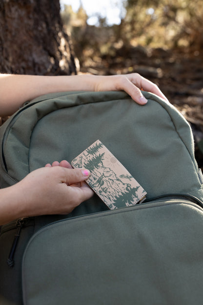 Close-up of hands tucking a slim, forest-inspired SD card organizer into the front zipper pocket of an olive green travel backpack.