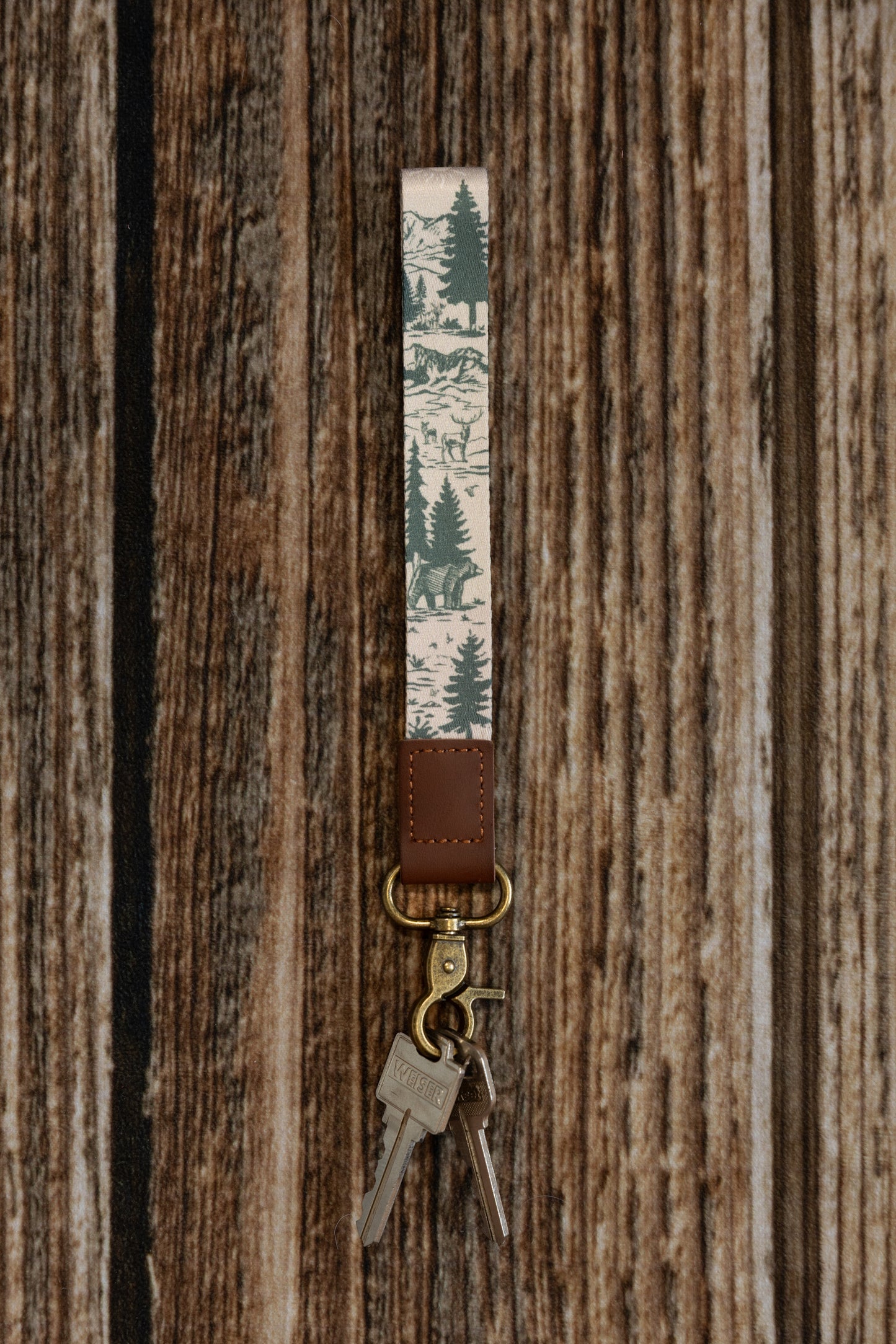 A vertical product shot of a forest-themed wristlet keychain against a rustic wood backdrop. The fabric displays an intricate mountain landscape with deer and bears, attached to a brown leather end and antique brass clasp holding silver keys.