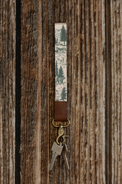 A vertical product shot of a forest-themed wristlet keychain against a rustic wood backdrop. The fabric displays an intricate mountain landscape with deer and bears, attached to a brown leather end and antique brass clasp holding silver keys.