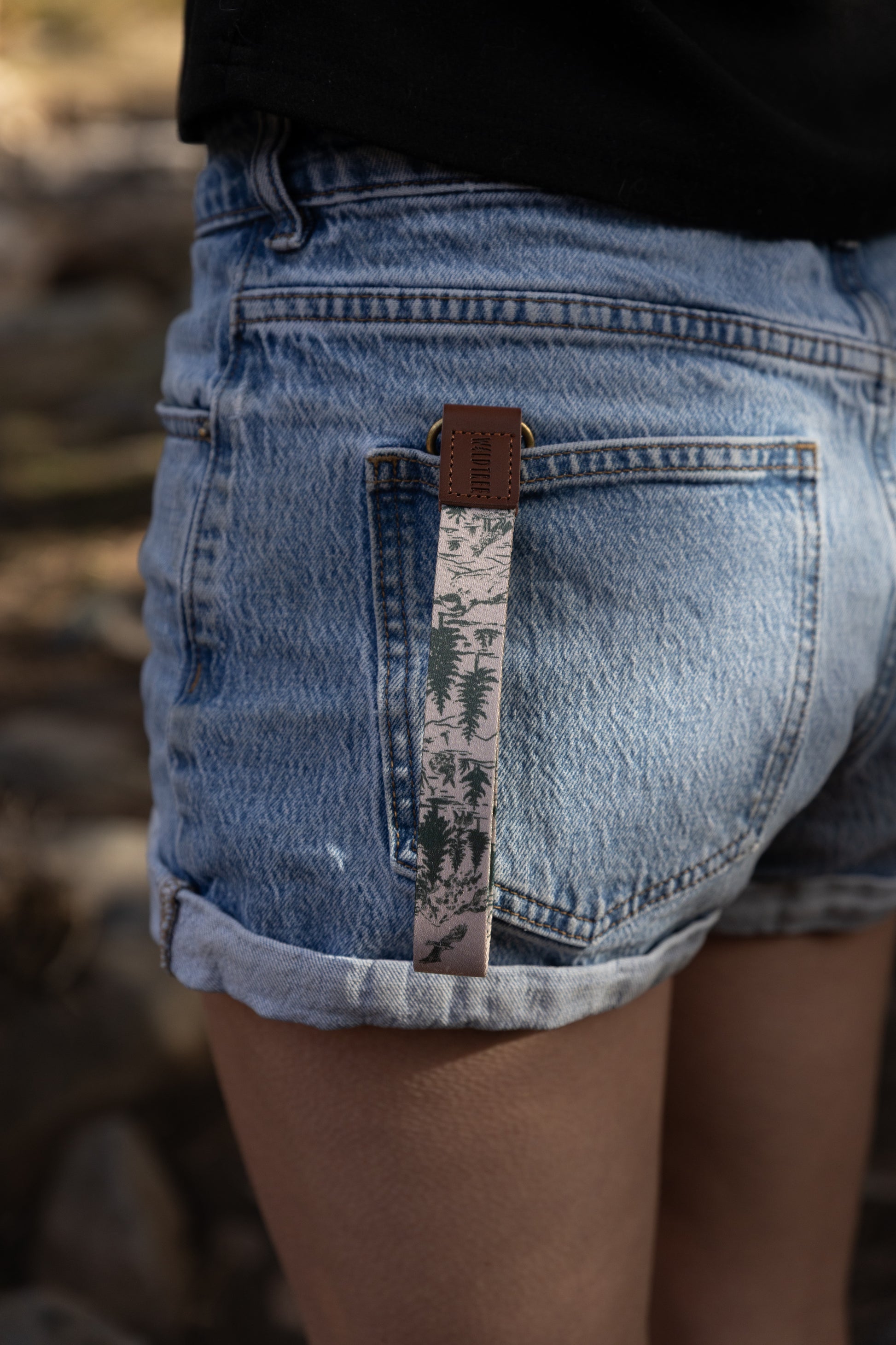 Lifestyle photography of the woodland forest key strap clipped to a denim pocket in a blurred outdoor setting. The design features soaring birds and mountain pines, emphasizing the rugged outdoor aesthetic of the WILDTREE brand.