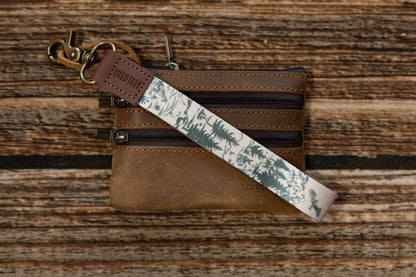 A mountain landscape wristlet keychain featuring eagles and evergreens resting on a brown leather multi-zipper wallet. The antique brass hardware and embossed WILDTREE leather tab add a rugged, high-quality feel to the outdoor-themed design.