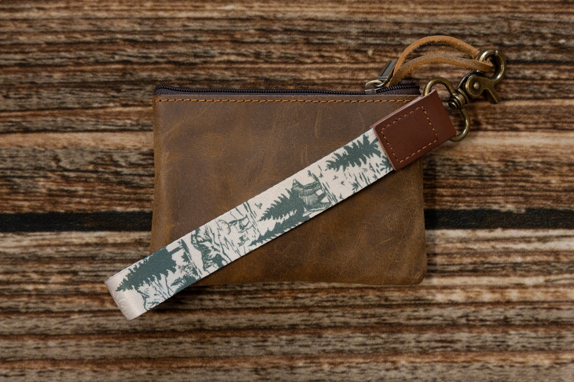A hand-drawn woodland fabric keychain laid diagonally across a distressed brown leather zipper pouch. The strap highlights a forest scene with a bear and pine trees, connected by a vegan leather end branded with WILDTREE.