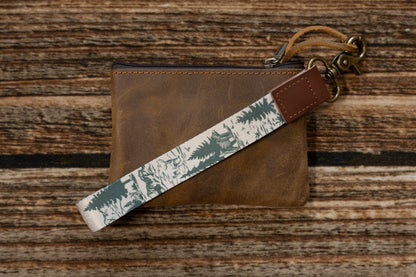 A hand-drawn woodland fabric keychain laid diagonally across a distressed brown leather zipper pouch. The strap highlights a forest scene with a bear and pine trees, connected by a vegan leather end branded with WILDTREE.