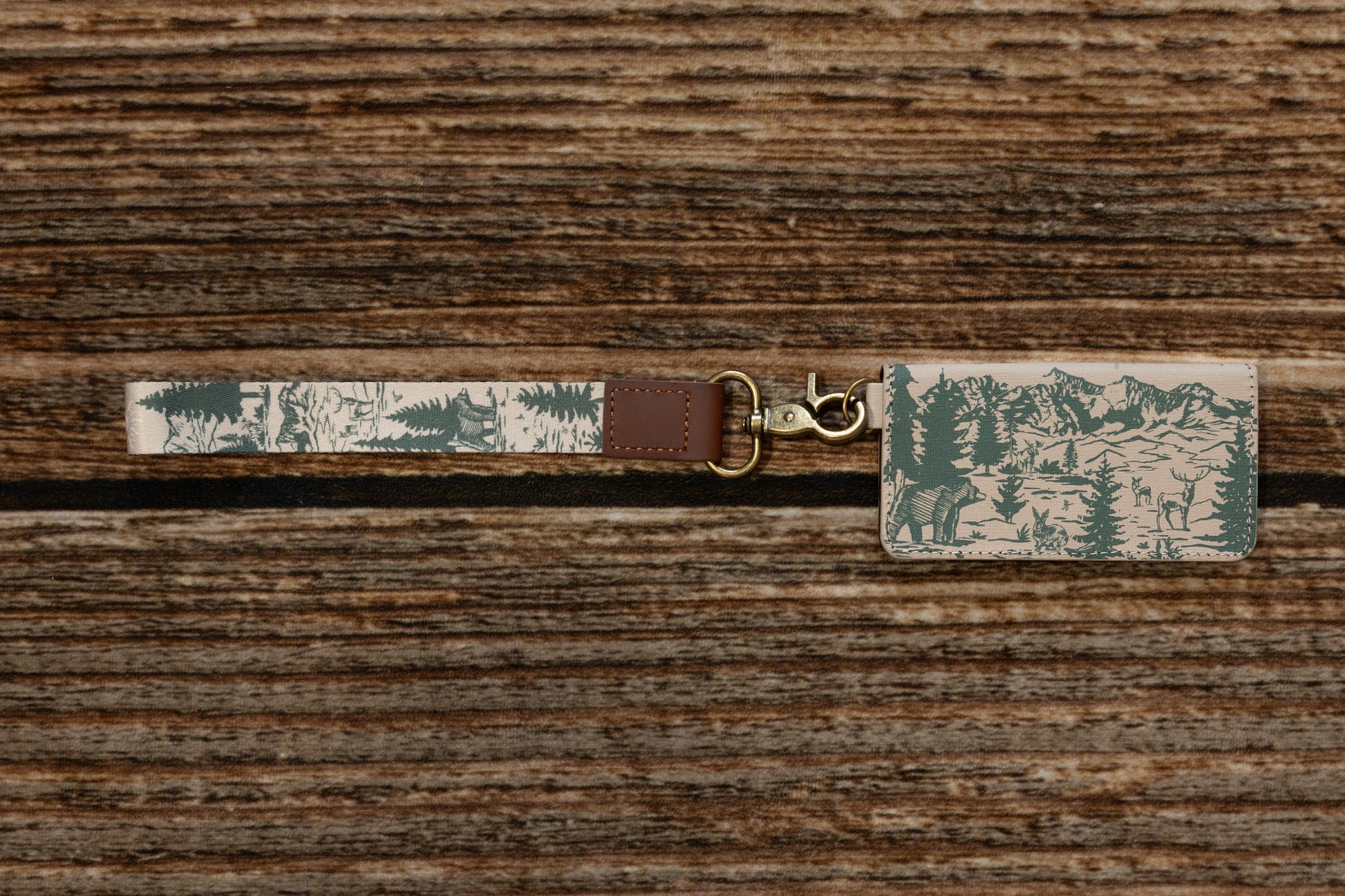 Top-down view of a nature-inspired key strap and matching patterned card holder on a dark wood background. The design shows a detailed green and beige mountain landscape with bears and evergreen trees, featuring a bronze swivel hook.