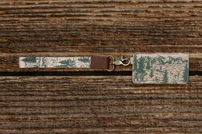 Top-down view of a nature-inspired key strap and matching patterned card holder on a dark wood background. The design shows a detailed green and beige mountain landscape with bears and evergreen trees, featuring a bronze swivel hook.