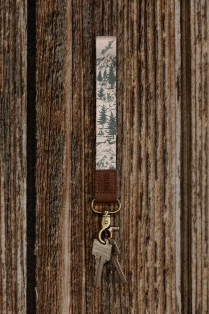 Close-up of a high-quality fabric wristlet keychain with a beige and green nature pattern. The hand-drawn illustrations include a rabbit, fox, and soaring eagle, finished with a sturdy brown leather end and bronze-finish hardware.