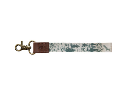 A clean product studio shot of the Woodland Landscape wristlet keychain isolated on a white background. The design features a hand-drawn forest pattern with evergreens and a soaring eagle, attached to a brown vegan leather tab embossed with the WILDTREE logo and a bronze antique-style swivel hook.