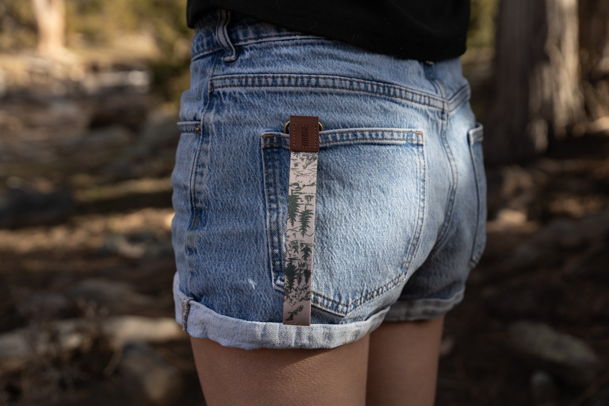 A close-up lifestyle shot of the Woodland Landscape wristlet keychain tucked into the pocket of light-wash denim shorts. The fabric strap features a green hand-drawn forest and eagle design, finished with a brown leather WILDTREE tab and antique brass hardware.