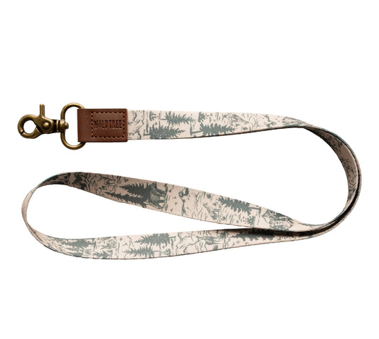 "Wildtree Woodland Landscape Neck Lanyard" on white. Tan strap with dark green pine trees and light gray mountain silhouettes. Brown leather loop ("WILDTREE") connects to a bronze swivel clasp. Curved to show the woodland print.