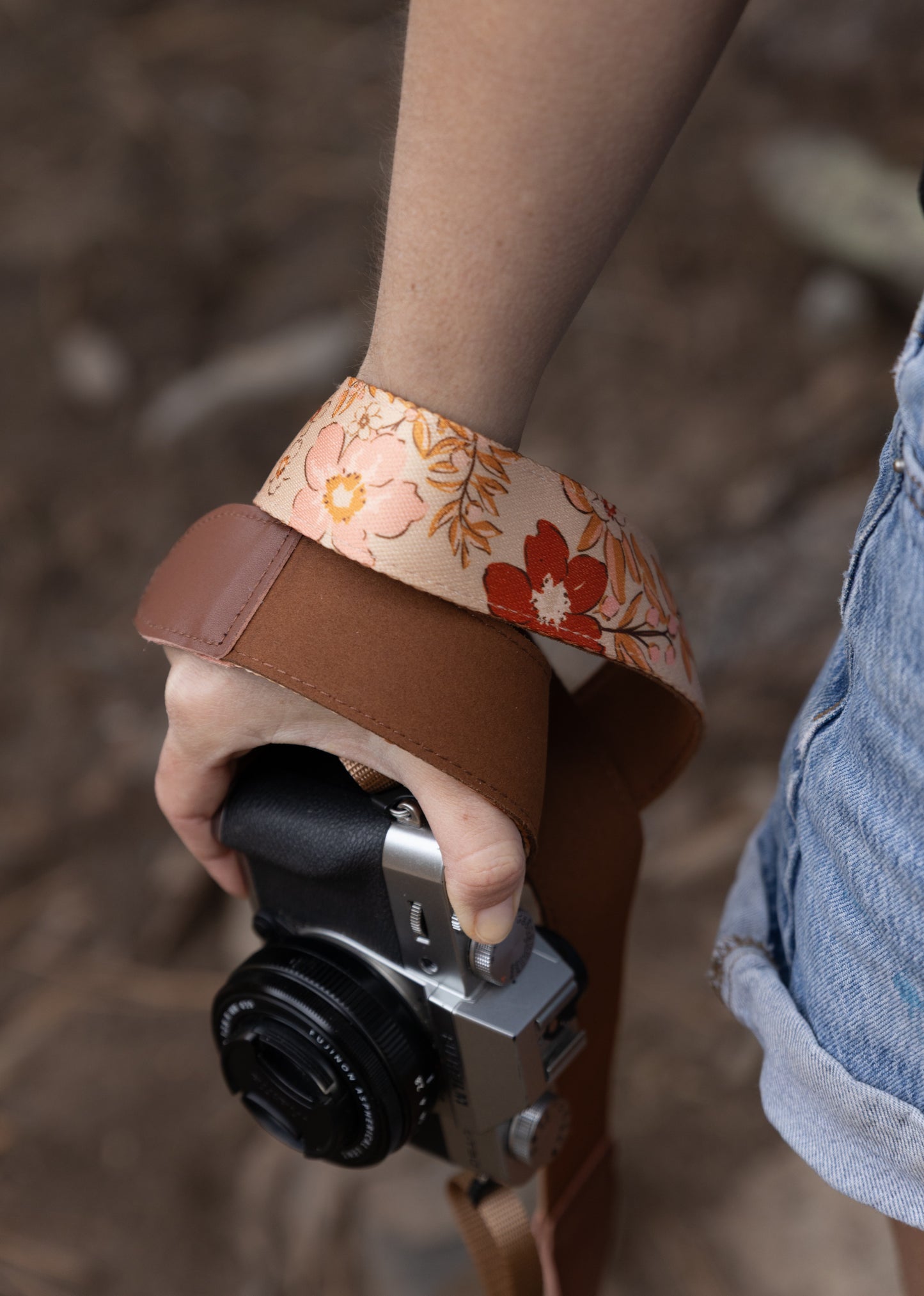 A close-up shot of a hand holding a silver and black mirrorless camera in a wooded setting. A "Mellow Meadow" camera strap, featuring a vintage-style floral pattern in shades of peach, cream, and rust, is wrapped securely around the person's wrist. The strap is accented with smooth brown leather ends.