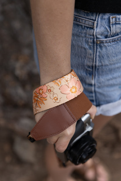 A vertical close-up of a person's wrist with the Mellow Meadow camera strap looped around it. The cream-colored fabric strap displays a detailed illustration of wildflowers and ferns. The person is wearing denim shorts and holding the camera body just out of focus at the bottom of the frame.