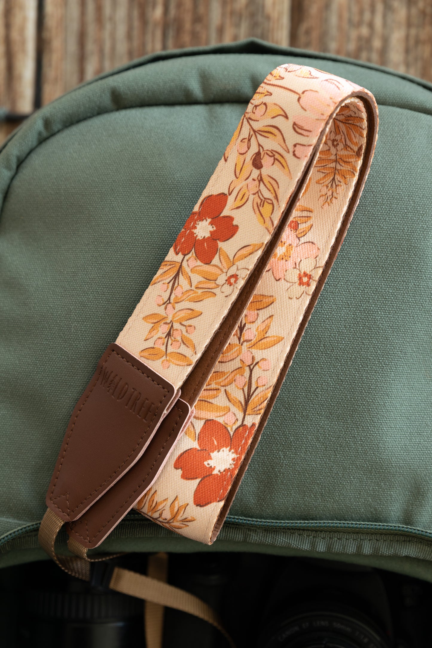 The floral camera strap is folded over itself on top of a green backpack. The focus is on the textured fabric weave of the cream, orange, and tan botanical pattern and the stitched brown leather ends.