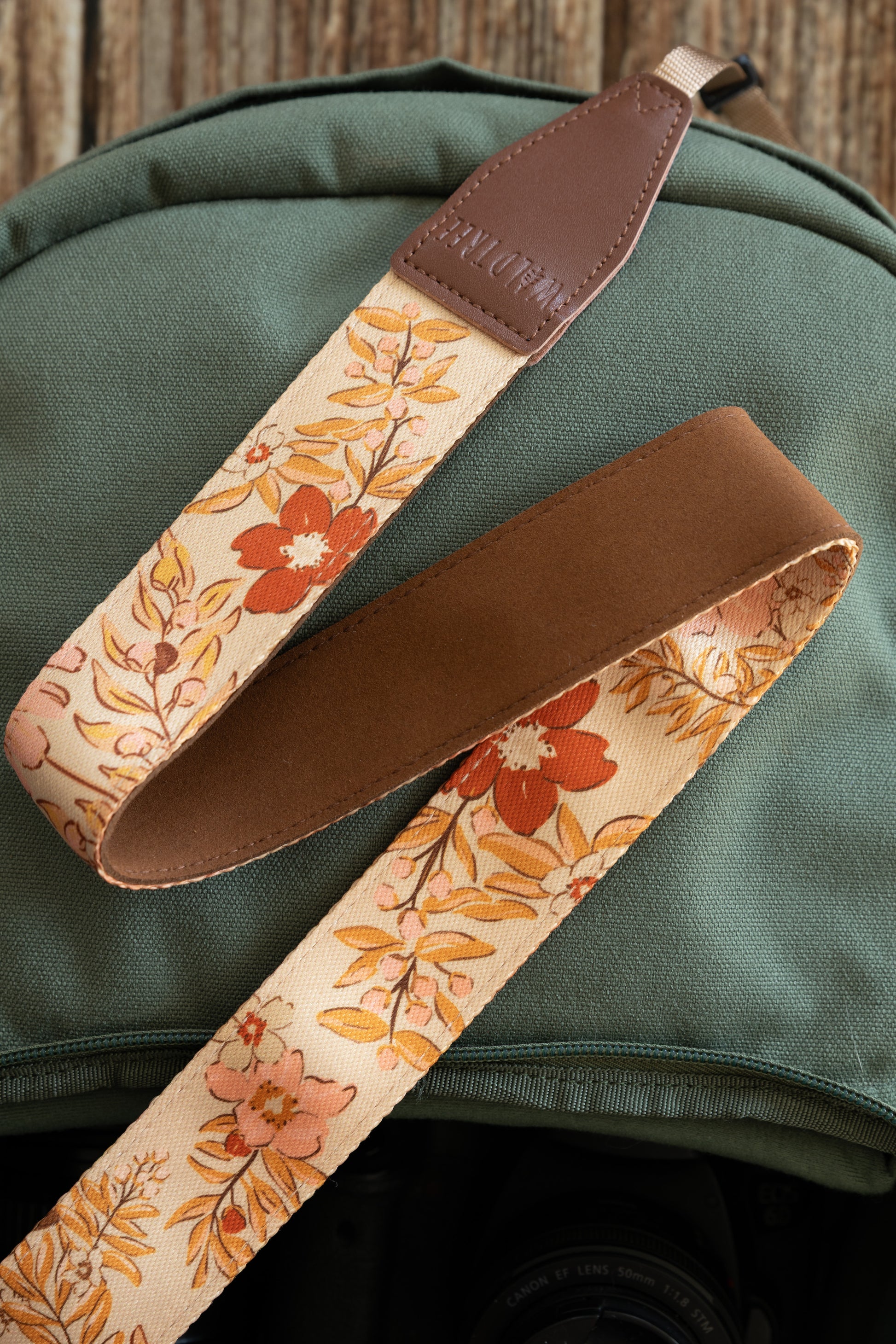A detailed view of the camera strap showing both the floral patterned side and the solid brown, non-slip underside. The strap is draped over a green camera bag, showing the high-quality stitching on the leather attachment.