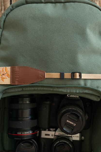 A close-up focusing on the adjustable tan nylon webbing and black plastic buckle of the camera strap. The strap is positioned near the open compartment of a green camera bag, where various lenses and camera bodies are visible.