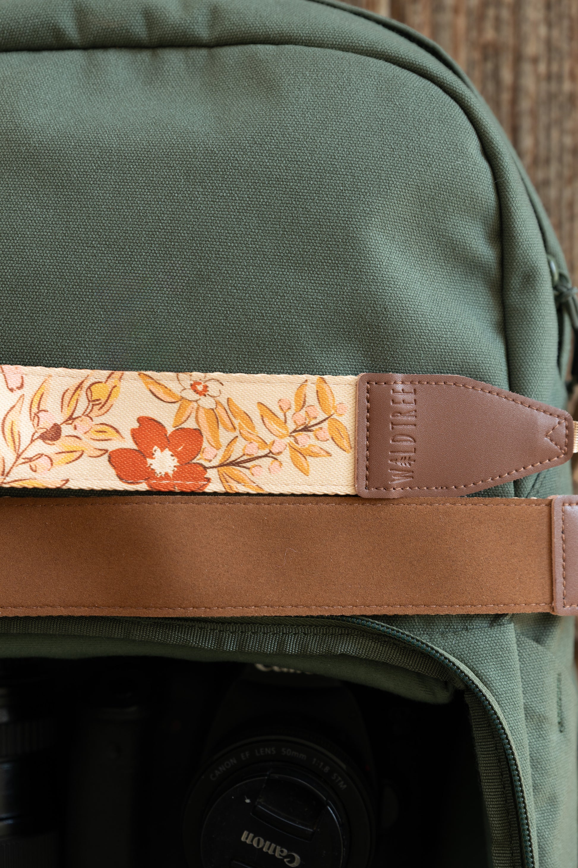 A close-up shot of the "Mellow Meadow" camera strap resting against a dark olive green canvas backpack. The image highlights the detailed floral pattern of the fabric, the brown leather connector piece, and the soft brown underside of the strap.