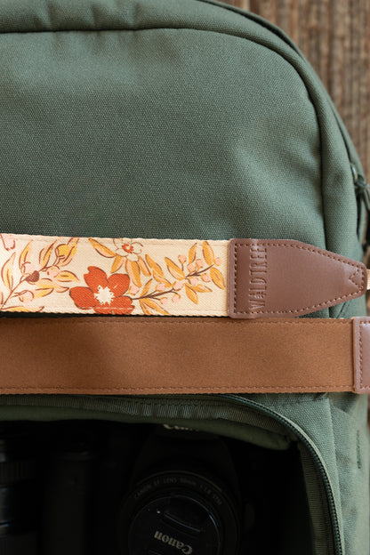 A close-up shot of the "Mellow Meadow" camera strap resting against a dark olive green canvas backpack. The image highlights the detailed floral pattern of the fabric, the brown leather connector piece, and the soft brown underside of the strap.