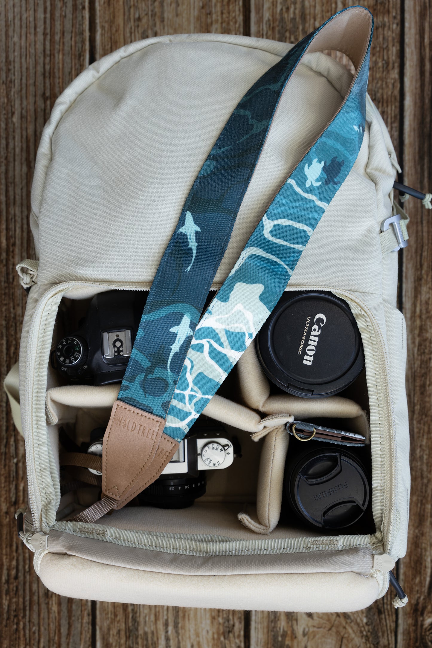 Top-down view inside a cream-colored camera bag showing a WILDTREE camera strap resting across the top, featuring a blue marine-themed print with sharks, sea turtles, and water-inspired patterns, placed above a camera body and lenses.