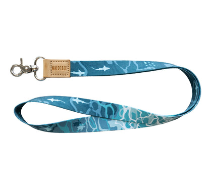 A blue and teal lanyard with an ocean-themed pattern of sharks and sea turtles is shown against a white background. It has a tan leather tab with "WILDTREE" embossed on it.