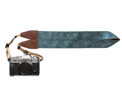 A product studio shot against a white background featuring a silver and black Fujifilm X-T30 mirrorless camera attached to a Wildtree "Shadow Garden" strap. The strap is extended to the right, showcasing its dark teal fabric printed with light grey botanical illustrations. The strap connects to the camera via brown leather ends and tan nylon webbing.