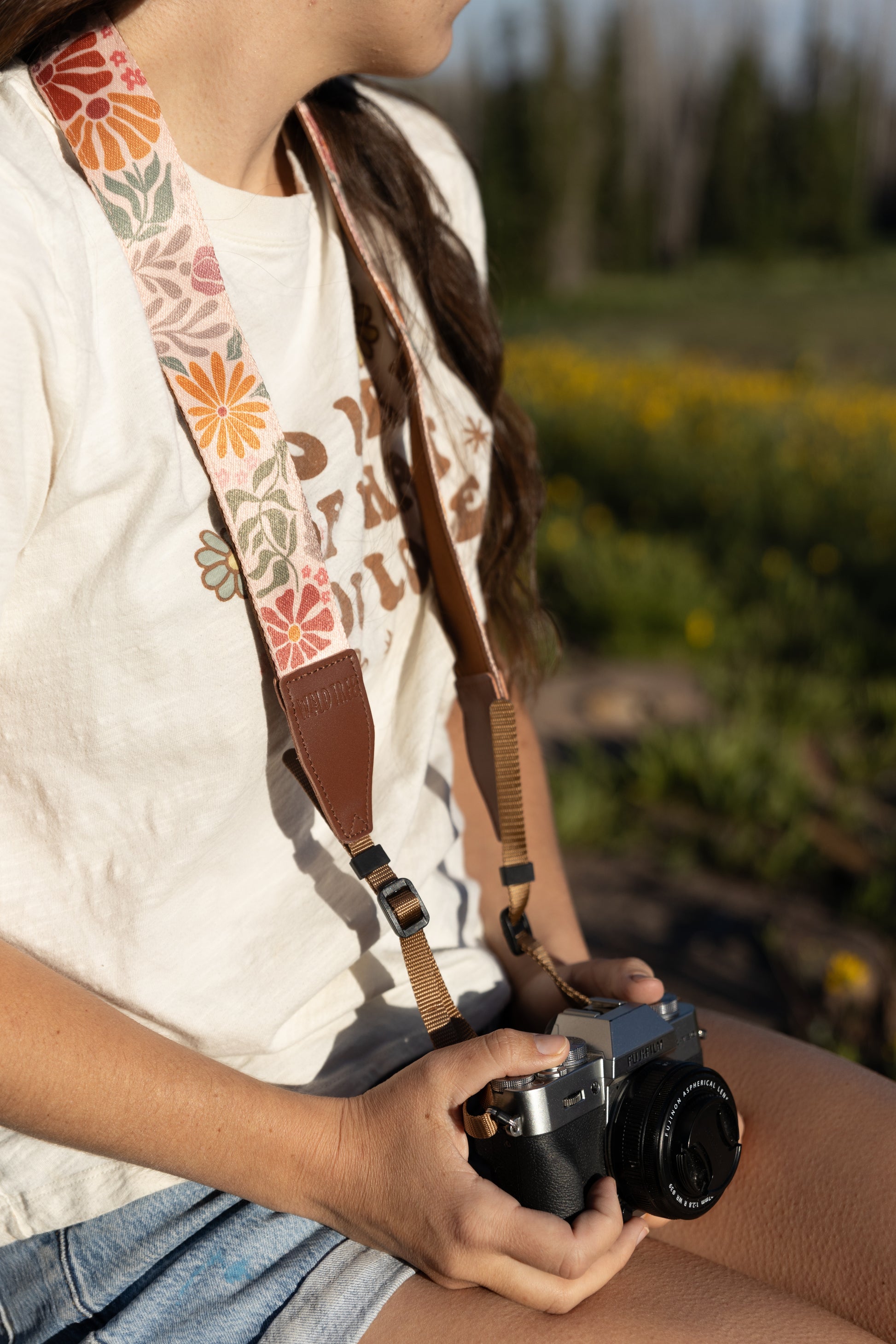 A close-up of a person's chest and hands, sitting down and holding a black and silver camera. They are wearing a cream-colored t-shirt with a graphic design. The camera strap is draped over their shoulder, displaying a sunset-colored floral design with large orange and pink daisies and green leaves on a light pink background.