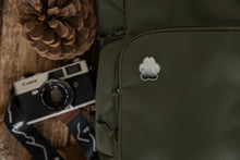 Load image into Gallery viewer, Acrylic pin in the shape of a paw designed with trees and mountains within the paw pinned to a green backpack with a camera and pinecone close by.
